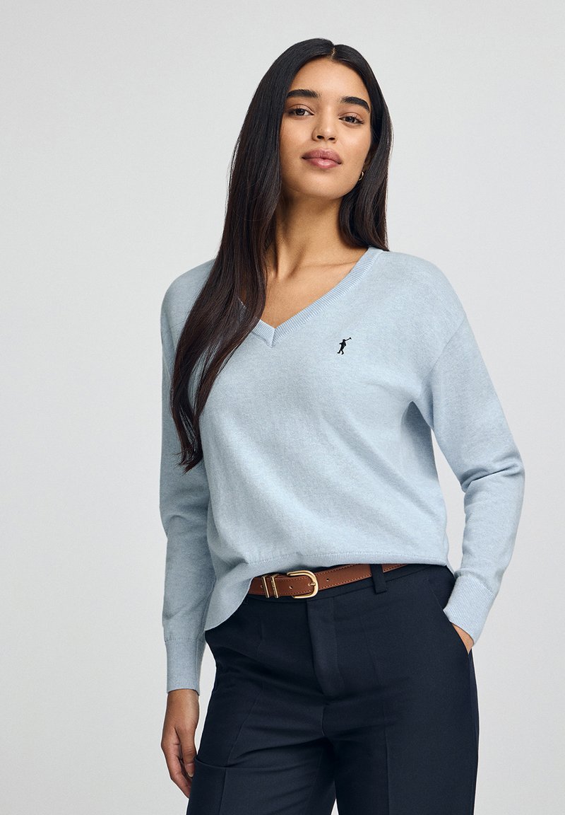 Ljusblå v-ringad tröja i mjuk tyg, med långa ärmar och ett litet svart emblem på bröstet, kombinerad med mörka byxor och ett brunt bälte.