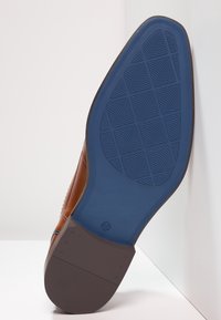 Brauner Lederschuh mit einer blauen Gummisohle, die ein gemustertes Profil und einen strukturierten Rand aufweist. Größe 42 ist auf der Sohle markiert.