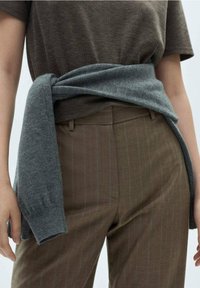 Grijze gebreide trui om de taille geknoopt over een bruin gestreept shirt en bruine broek. De materialen lijken zacht met een ontspannen pasvorm.