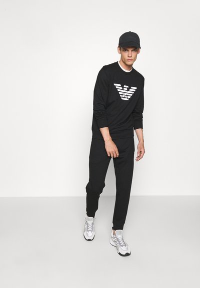 Juodas džemperis su baltu dryžuotu logotipu, derinamas su juodomis kelnėmis ir baltais sportiniais bateliais. Apranga pasižymi kasdieniu siluetu ir patogiu dizainu.