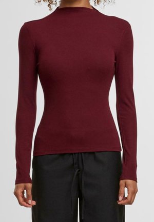 Personne portant un haut bordeaux cintré à manches longues avec un col rond haut et un pantalon noir à cordon, debout devant un fond uni.