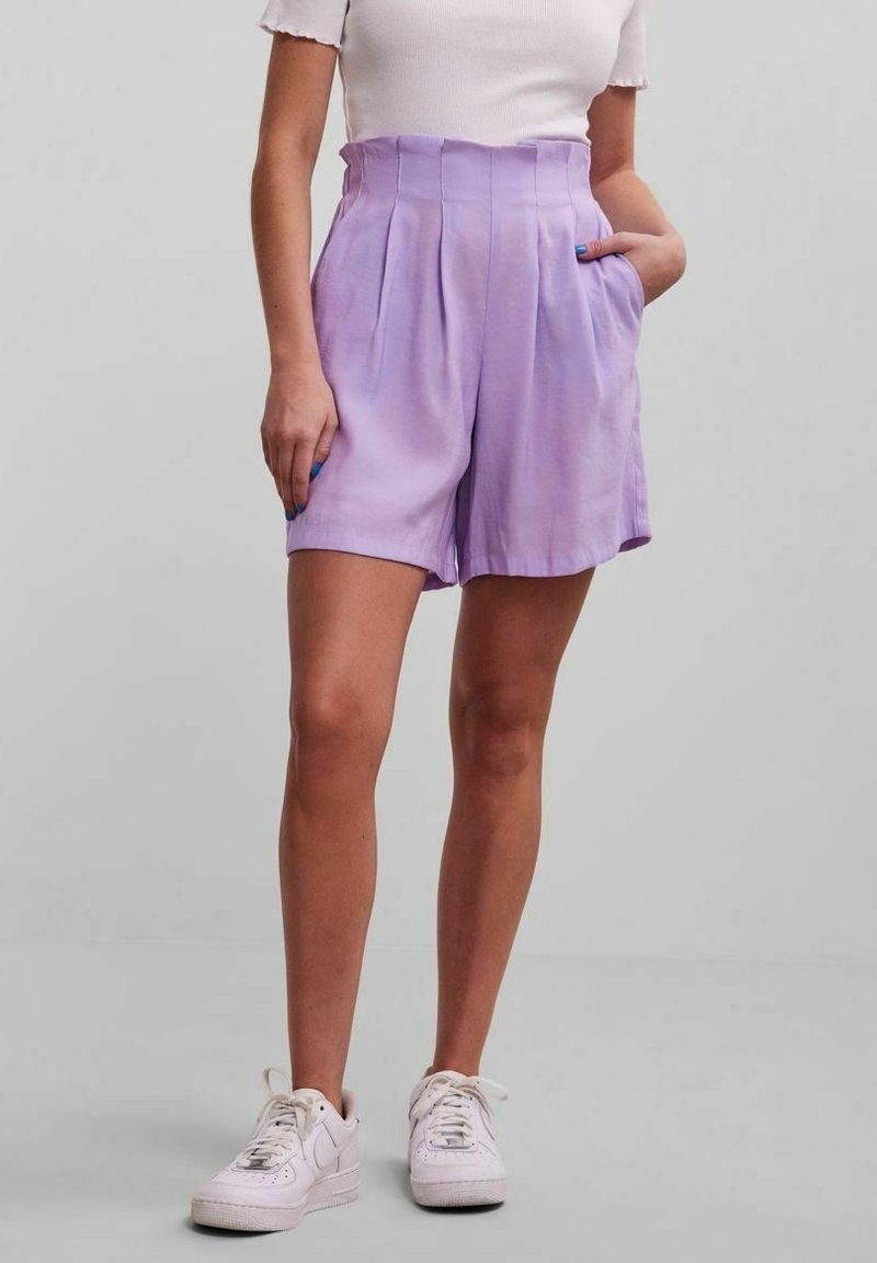 Lavendelfärgade plisserade shorts i mjukt tyg, med hög midja, sidofickor och en avslappnad passform, matchade med vita sneakers.