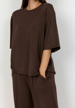 Femme portant un haut marron ample à manches courtes et un pantalon assorti, une main dans la poche, sur un fond clair uni.