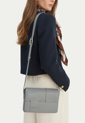 Žena v tmavě modrém saku a vzorovaném hedvábném šátku nese světle šedou kabelku Gino Rossi přes tělo.