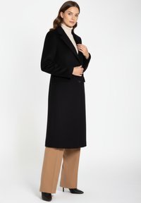 Zwarte lange jas met inzettende revers en een enkele knoopsluiting, gecombineerd met beige wijde broekspijpen en zwarte pumps met puntige neus.