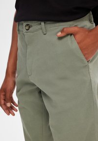 Pantalones de algodón verde claro con un bolsillo frontal visible y cierre de botón. La tela tiene una textura suave y el diseño presenta un ajuste recto.