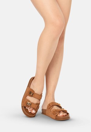 Piernas con sandalias marrones de ante sin cordones con dos correas ajustables con hebilla, de pie sobre un fondo claro y liso.