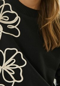 Pull noir avec un col côtelé et un détail de broderie florale blanche. Texture douce, coupe ajustée et accents décoratifs prononcés.