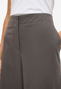 Vyötäröltä korkeat, väljät harmaat housut, jotka on tehty sileästä kankaasta, sivutaskuilla ja piilotetulla vetoketjulla, esitellen sulavan, saumattoman muotoilun.