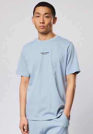 Junger Mann trägt hellblaues kurzärmeliges T-Shirt und passende Hose, steht mit einer Hand in der Tasche, neutraler Ausdruck, einfarbiger grauer Hintergrund.