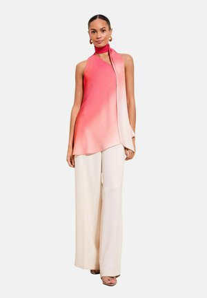 Korallrózsaszín nyakpántos felsőt viselő nő, amelyhez sál kapcsolódik, fehér, bő szárú nadrágban, fehér háttér előtt áll.