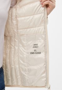 Lys beige polstret jakke med en glat tekstur, quiltet design og "Dyrevenlig" mærkater, der angiver 0% dunfjer.