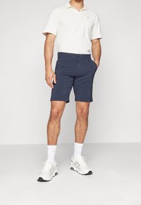 Marineblå shorts med flad frontdesign, lavet af letvægtsstof. Båret med en cremefarvet polo og hvide sneakers med grå detaljer.