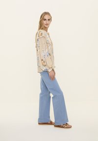 Blouse à manches longues, de couleur beige clair, avec des détails froncés, associée à un jean en denim bleu clair à pattes d'éléphant et des sandales marron avec une boucle.