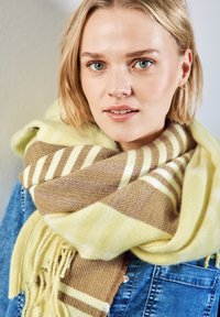 Beige en gele gestreepte sjaal met franjes, gemaakt van zacht materiaal, om de nek gewikkeld, gelaagd over een spijkerjack.