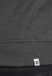 Dark grey sweatshirt featuring a ribbed hem, smooth texture, and a small white label with black logo at the bottom left corner.