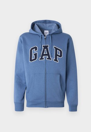 Hanorac albastru cu fermoar, buzunare frontale, glugă cu șnur și litere mari „GAP” pe piept, în nuanțe de bleumarin și alb.