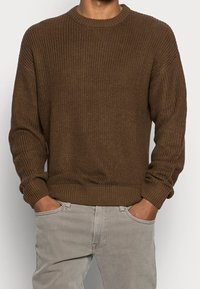 Brun ribbad stickad tröja med rund halsringning och långa ärmar, som har en texturerad yta och en åtsittande midja. I kombination med ljusgrå byxor.