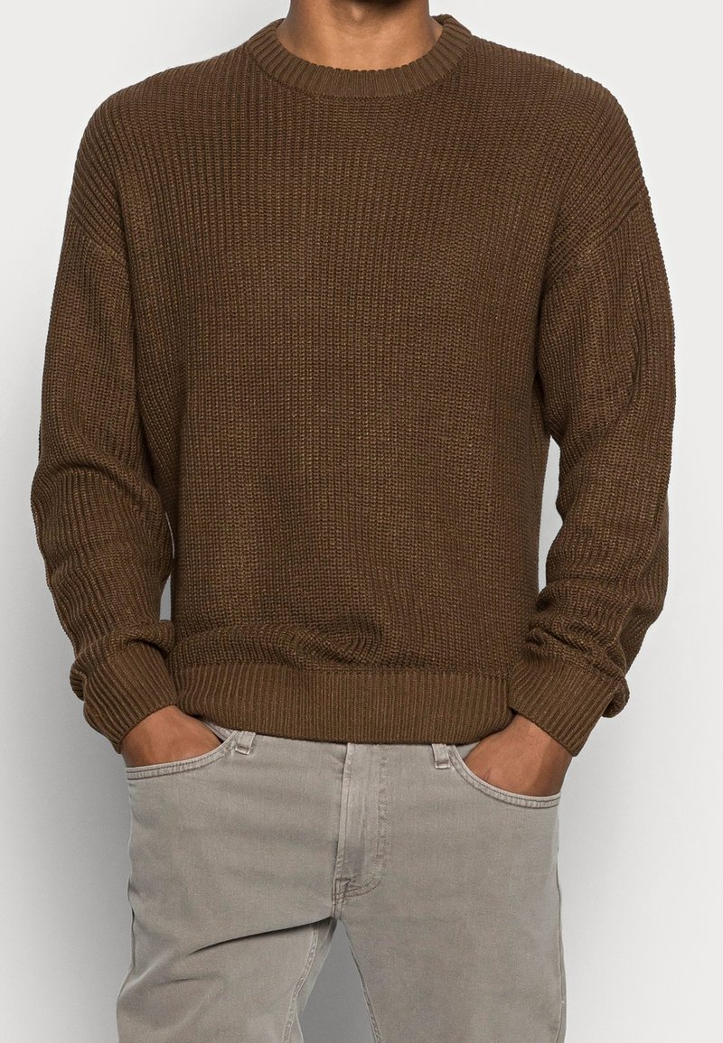 Brun ribbad stickad tröja med rund halsringning och långa ärmar, som har en texturerad yta och en åtsittande midja. I kombination med ljusgrå byxor.