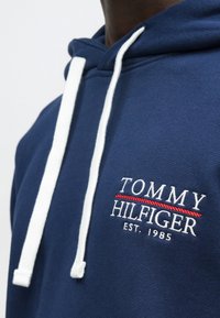 Κοντινή λήψη σκούρου μπλε φούτερ με κουκούλα, με λευκά κορδόνια και λευκό και κόκκινο κεντημένο λογότυπο "Tommy Hilfiger Est. 1985" στο στήθος.