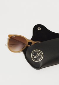 Ochelari de soare cu lentile degradate maro, ramă mată de culoare cămilă și logo-ul Ray-Ban, plasati parțial într-un etui din piele neagră cu un emblemă aurie.