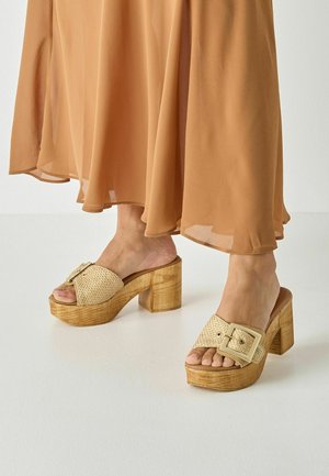 Beige gewebte Plateau-Mules mit einem breiten Riemen und einer quadratischen Schnalle, ergänzt durch einen klobigen Holzabsatz. Glatte Textur überall.