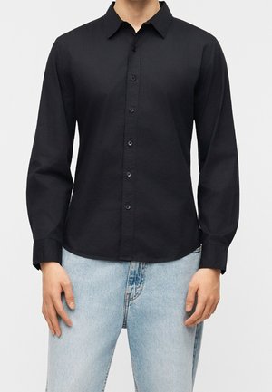 Homme portant une chemise noire ajustée à manches longues avec boutons et un jean bleu clair, debout devant un fond uni.