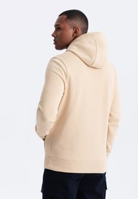 Felpa beige con un design aderente, dotata di cappuccio e polsini e orlo a coste. Il materiale appare morbido e testurizzato, indossata da una persona vista da dietro.
