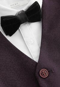 Gilet in tessuto di un viola scuro con una camicia bianca sotto, caratterizzato da un papillon in velluto nero e un bottone bordeaux.