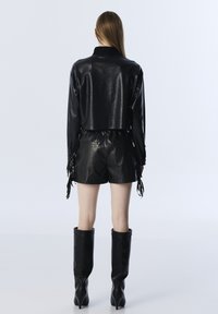 Korte zwarte leren jas met lange mouwen en een geribbelde kraag, gecombineerd met zwarte leren shorts met franjes aan de zijkant. Zwarte kniehoge laarzen.