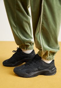 Svarta sportiga sneakers med texturerat tyg och nät, med rundad form och flexibel sula, tillsammans med gröna sweatpants med elastisk mudd.