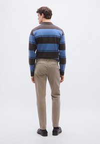 Ριγέ μακρυμάνικο πόλο σε καφέ και μπλε, συνδυασμένο με ανοιχτό μπεζ παντελόνι. Το ύφασμα φαίνεται απαλό· εφαρμοστή γραμμή· ομαλή υφή· φορεμένο με μαύρα παπούτσια.