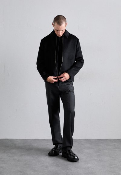 Black zip-up jacket with a collar, worn over a fitted black shirt, paired with slim gray trousers and black shoes on a gray floor backdrop.
