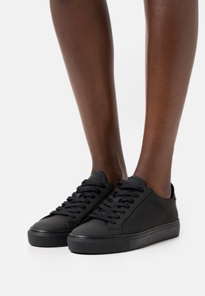 Ben iført lavtop, helt sorte sneakers med tyk sål og sorte snørebånd, stående på en hvid overflade med en hvid baggrund.