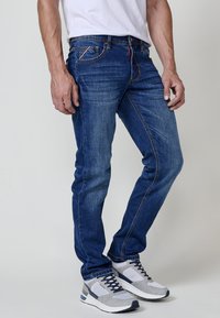 Vaqueros de mezclilla azul con corte recto, con costuras naranjas y un ligero desvanecimiento. Combinados con zapatillas deportivas grises y blancas.