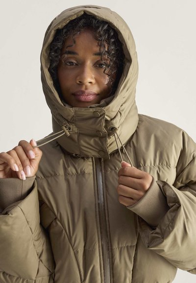 Mujer ajustándose la capucha de una chaqueta de invierno acolchada marrón con agujeros para el pulgar en las mangas, sobre un fondo claro y liso.