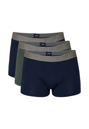 Mann steht barfuß in marineblauen Boxershorts mit grauem Bund vor einem schlichten weißen Hintergrund.