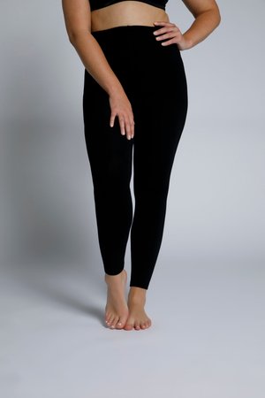 Frau steht barfuß, trägt schwarze hoch taillierte Leggings und ein schwarzes kurzes Oberteil, posiert mit einer Hand in der Hüfte und einer Hand, die auf dem Oberschenkel ruht.