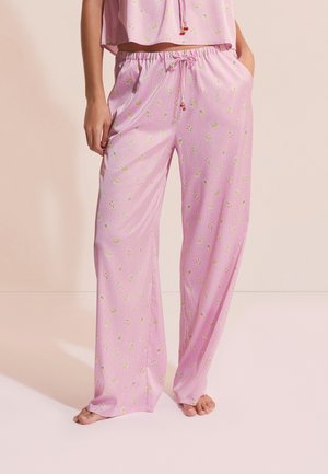 Luźne różowe spodnie od piżamy z drobnym kwiatowym nadrukiem i wiązaniem w pasie, noszone z dopasowaną różową bluzką, boso na jasnym tle.