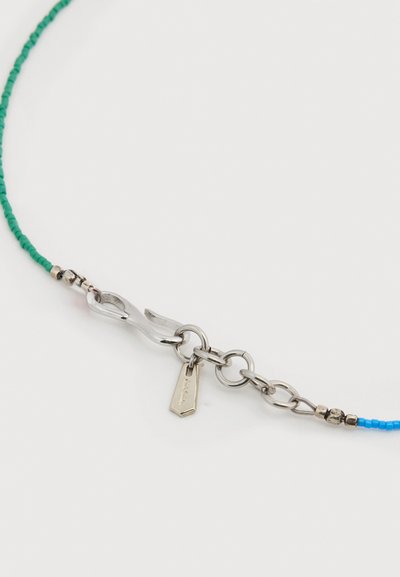 Ασημένιο κούμπωμα γαμπρών που συνδέει μπλε και πράσινες χάντρες με μικρή μεταλλική ετικέτα σε λευκό φόντο.