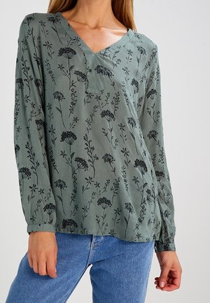 Femme portant un chemisier vert sauge à col en V avec des imprimés floraux noirs et un jean bleu, debout devant un fond neutre.