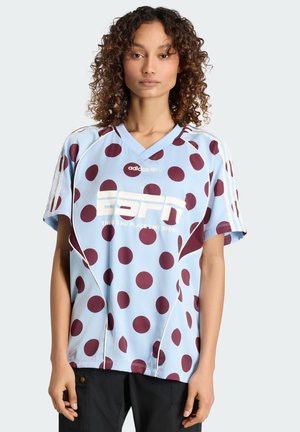 Mujer con cabello rizado, vistiendo una camiseta azul claro de Adidas con lunares granate, logo de ESPN y pantalones negros, sobre un fondo liso.