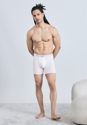 Άνδρας με dreadlocks, όρθιος και ξυπόλητος, φοράει λευκά μποξεράκια σε ένα φωτεινό, μινιμαλιστικό δωμάτιο κοντά σε μια καρέκλα με υφή.