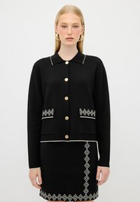 Svart cardigan med krage, guldknappar och två framfickor med ett vitt diamantmönster. Strukturerad tyg, matchad med en kjol.