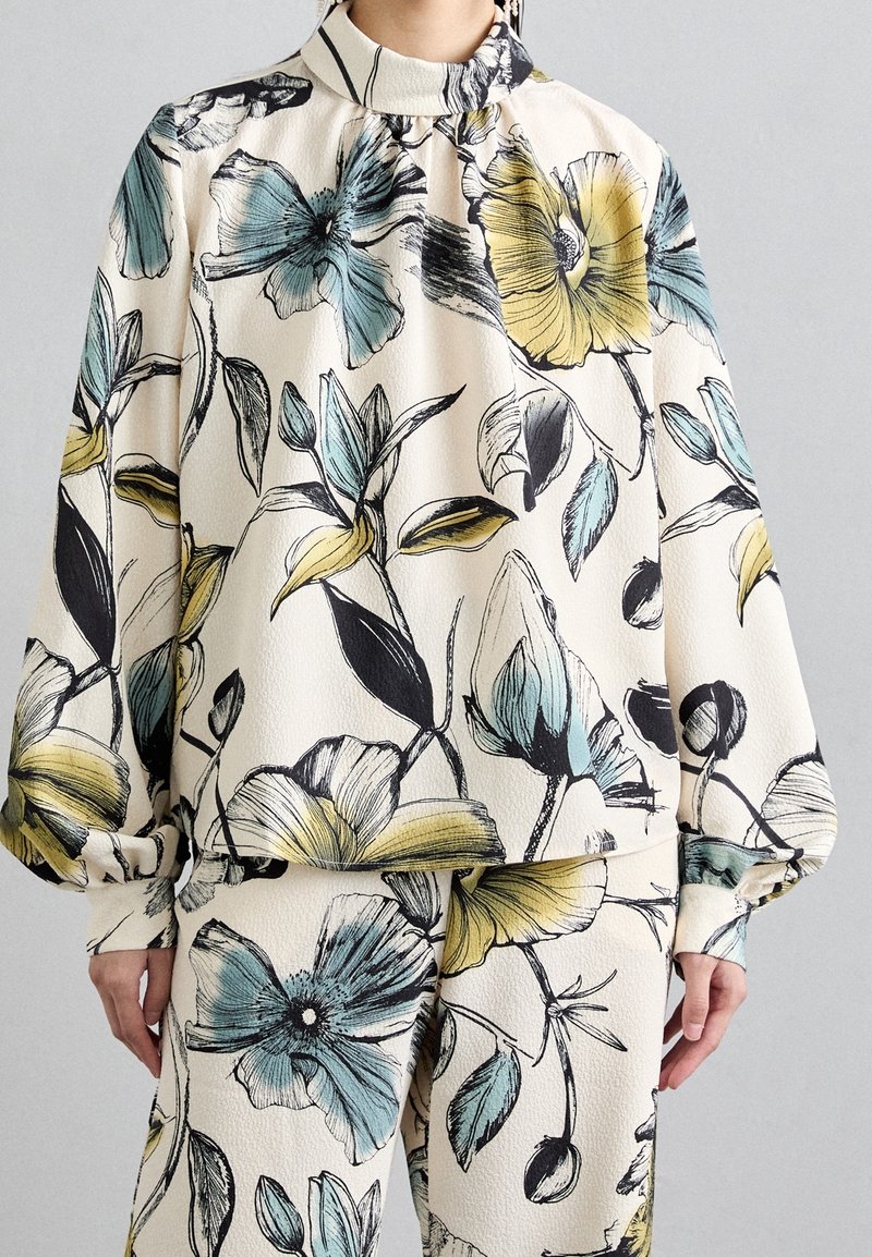 Haut et pantalon à motif floral crème, ornés de grandes fleurs bleues et jaunes. Tissu texturé avec un col montant et des détails froncés.