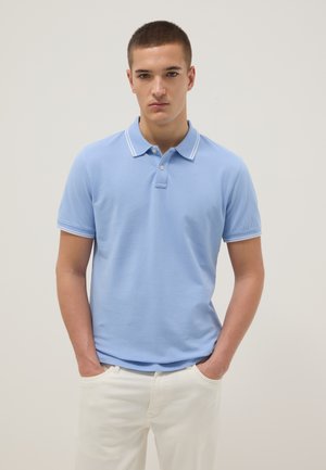 Jonge man met kort haar, gekleed in een lichtblauw poloshirt en witte broek, staand met handen in de zakken tegen een effen achtergrond.