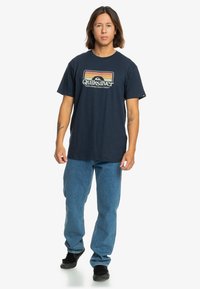Navyblaue Baumwoll-T-Shirt mit retro Quiksilver-Logo in Gelb, Orange und Weiß. Kombiniert mit hellblauen Jeans und schwarzen Schuhen.
