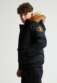 Giacca a vento nera imbottita con cappuccio foderato in pelliccia, dotata di una patch con logo sulla spalla, chiusura a zip frontale e tasche laterali. Texture liscia e design robusto.