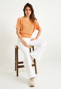 Un suéter de punto color naranja claro con mangas cortas, combinado con unos pantalones vaqueros acampanados blancos y zapatillas beige, sentado en un taburete de madera.