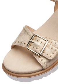 Sandale en daim beige avec des détails à clous dorés, une grande boucle dorée sur la bride avant et une semelle rembourrée.